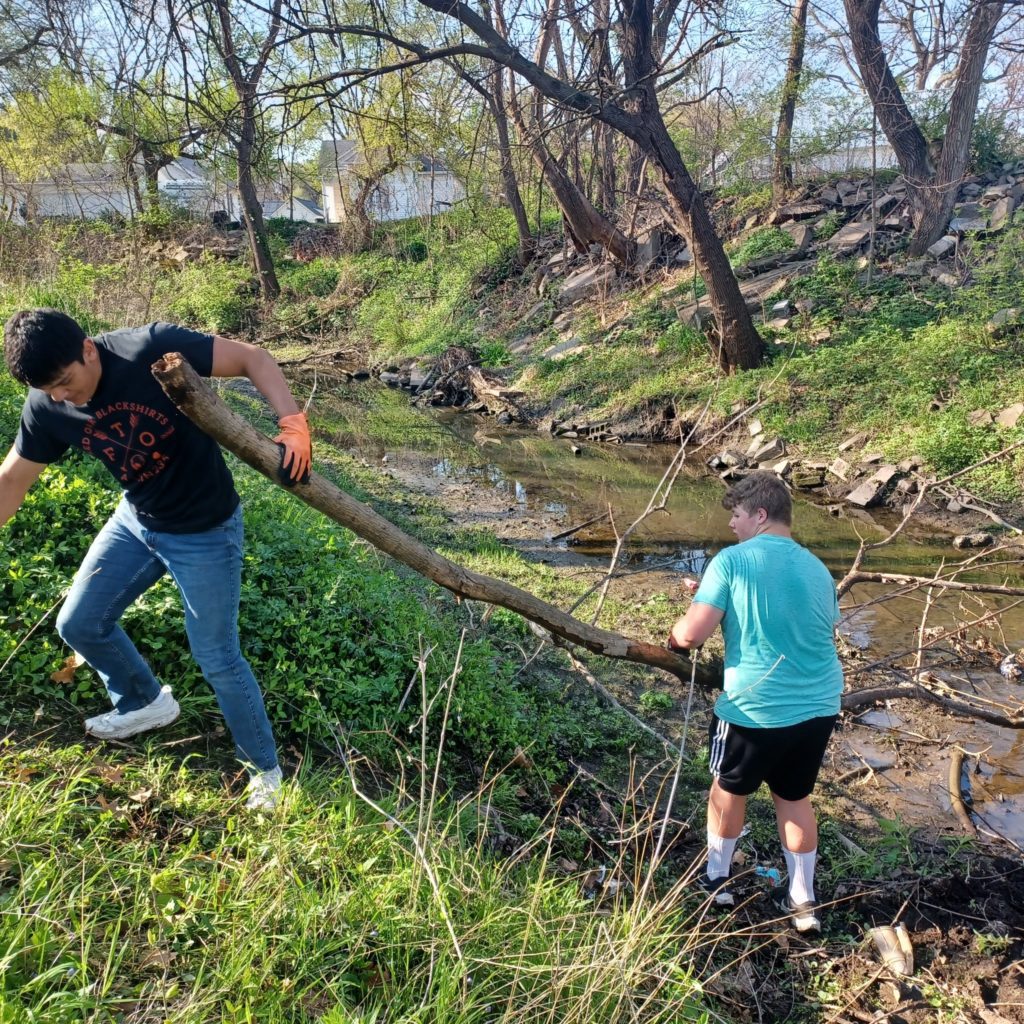 A kid helping another kid with a log while cleaning up a creek