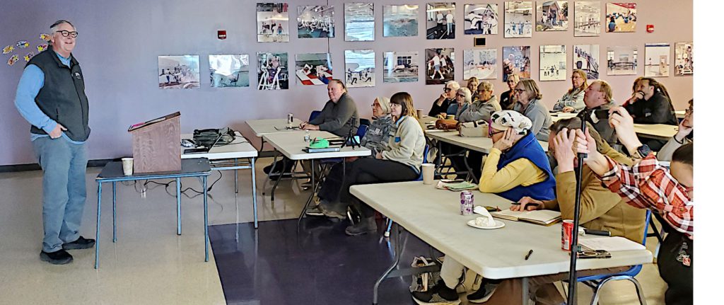 A man in a blue shirt with a black sleeveless vest speaks to a crowd in a meeting room.